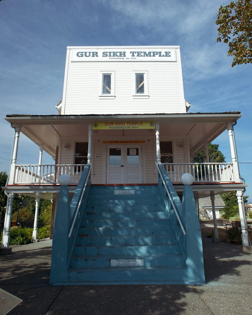 White false-front 3-storey building with blue concrete stairs to the front door on the second storey. Sign on the top of the false front reads White false-front 3-storey building with blue concrete stairs to the front door on the second storey. Sign on the top of the false front reads "Gur Sikh Temple Founded in 1911". 