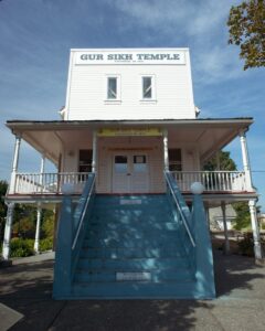 White false-front 3-storey building with blue concrete stairs to the front door on the second storey. Sign on the top of the false front reads "Gur Sikh Temple built 1911".